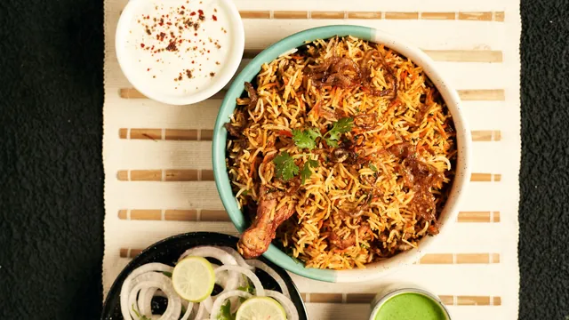 A bowl of chicken biryani with raita, onions and lime