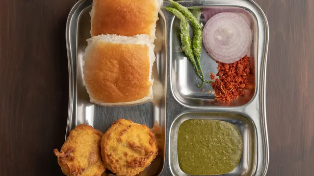 Vada pav served on a steel plate with chutneys, onion and green chillies